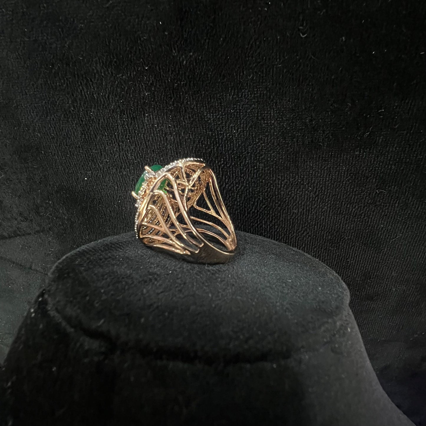 Close-up of an elegant statement ring featuring a large oval green gemstone at the center, surrounded by concentric circles of sparkling white diamonds set in a gold base against a black background.