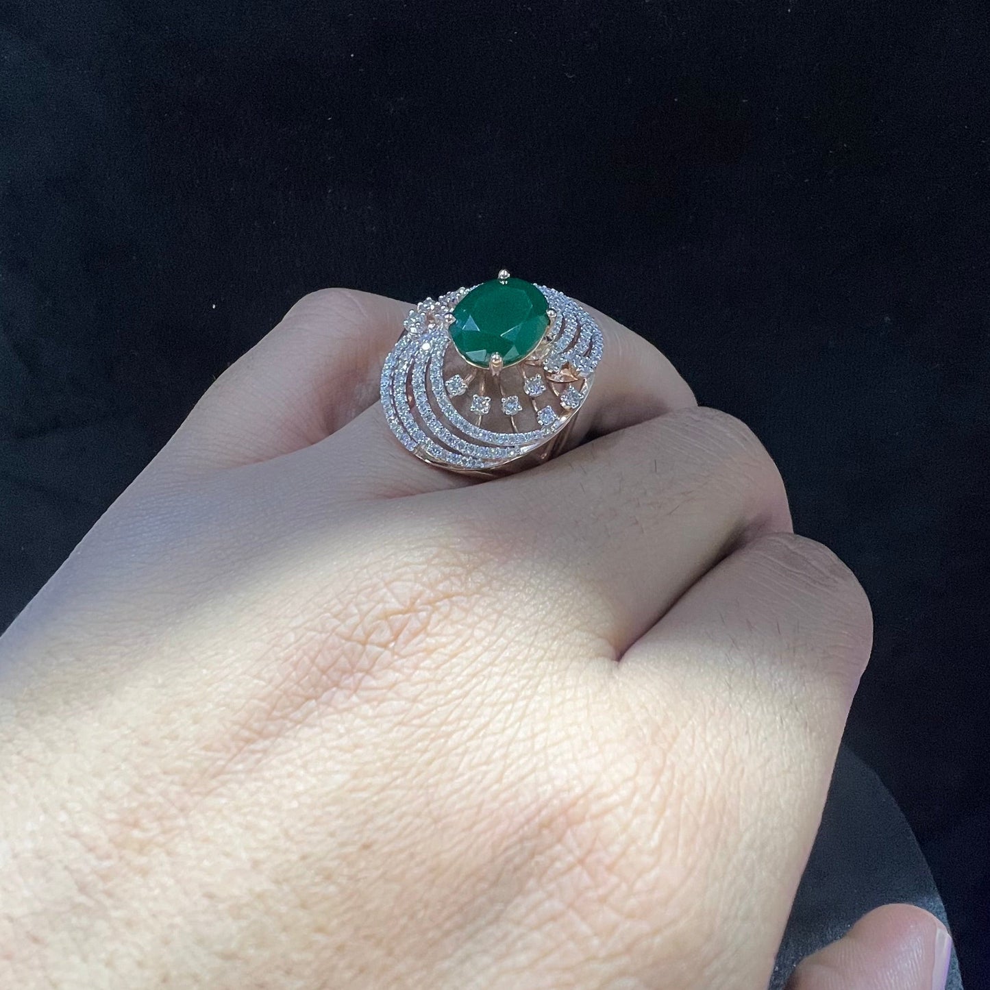 Close-up of a hand holding an elegant statement ring featuring a large oval green gemstone at the center, surrounded by concentric circles of sparkling white diamonds set in a gold base against a black background.
