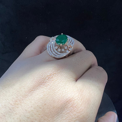 Close-up of a hand holding an elegant statement ring featuring a large oval green gemstone at the center, surrounded by concentric circles of sparkling white diamonds set in a gold base against a black background.