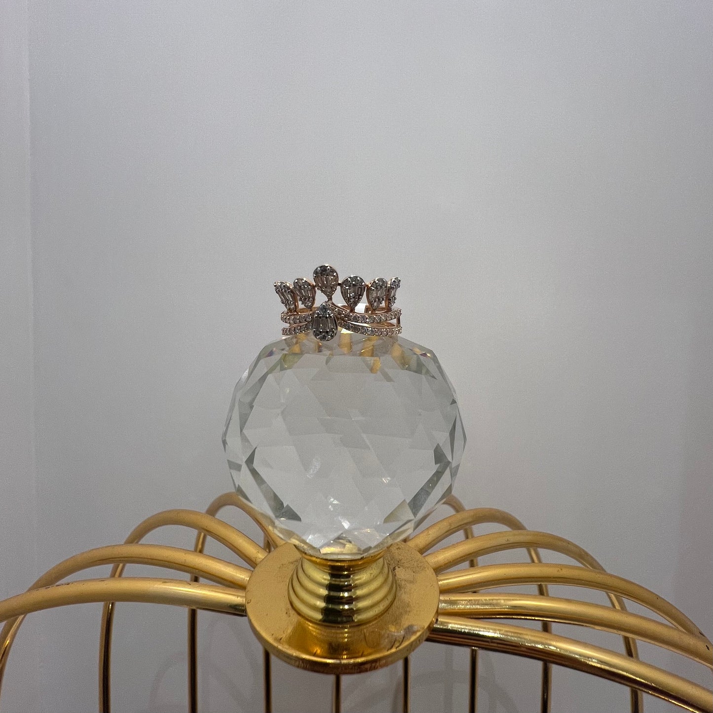 Close-up of a crown-inspired diamond ring featuring multiple baguette-shaped diamonds set upright along the top, paired with a pavé-set band in gold, displayed a glass ball that is on a gold color cage.