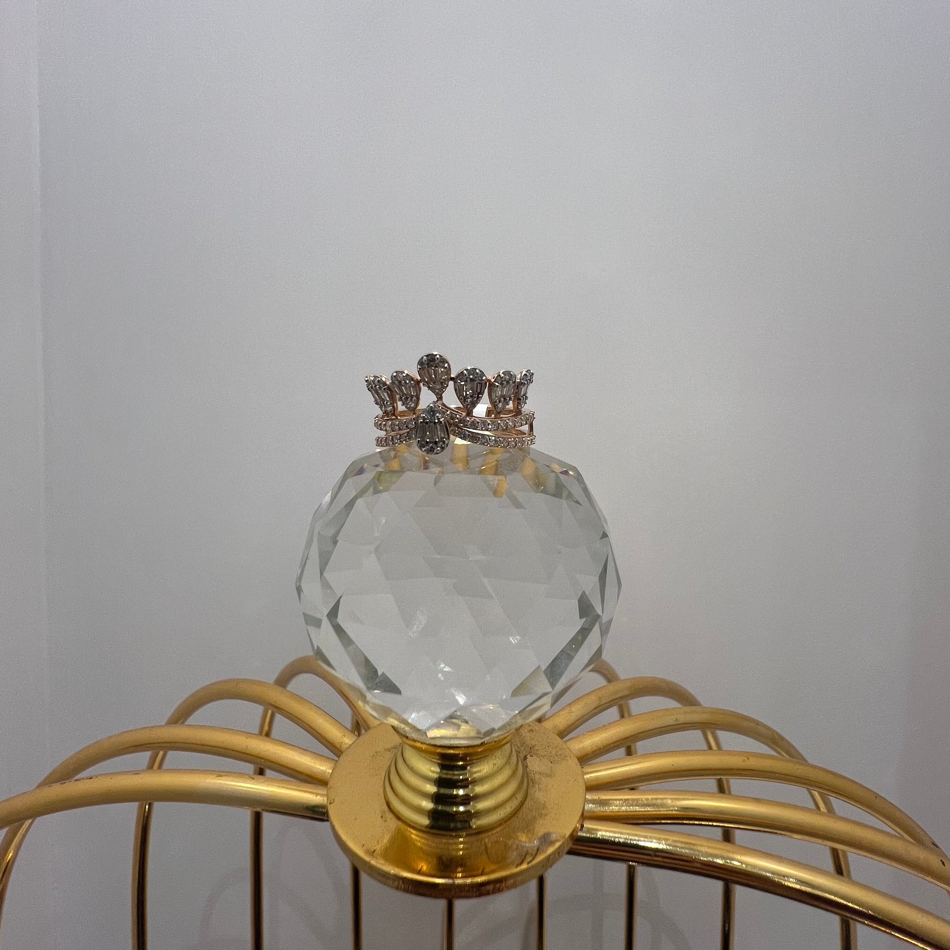 Close-up of a crown-inspired diamond ring featuring multiple baguette-shaped diamonds set upright along the top, paired with a pavé-set band in gold, displayed a glass ball that is on a gold color cage.