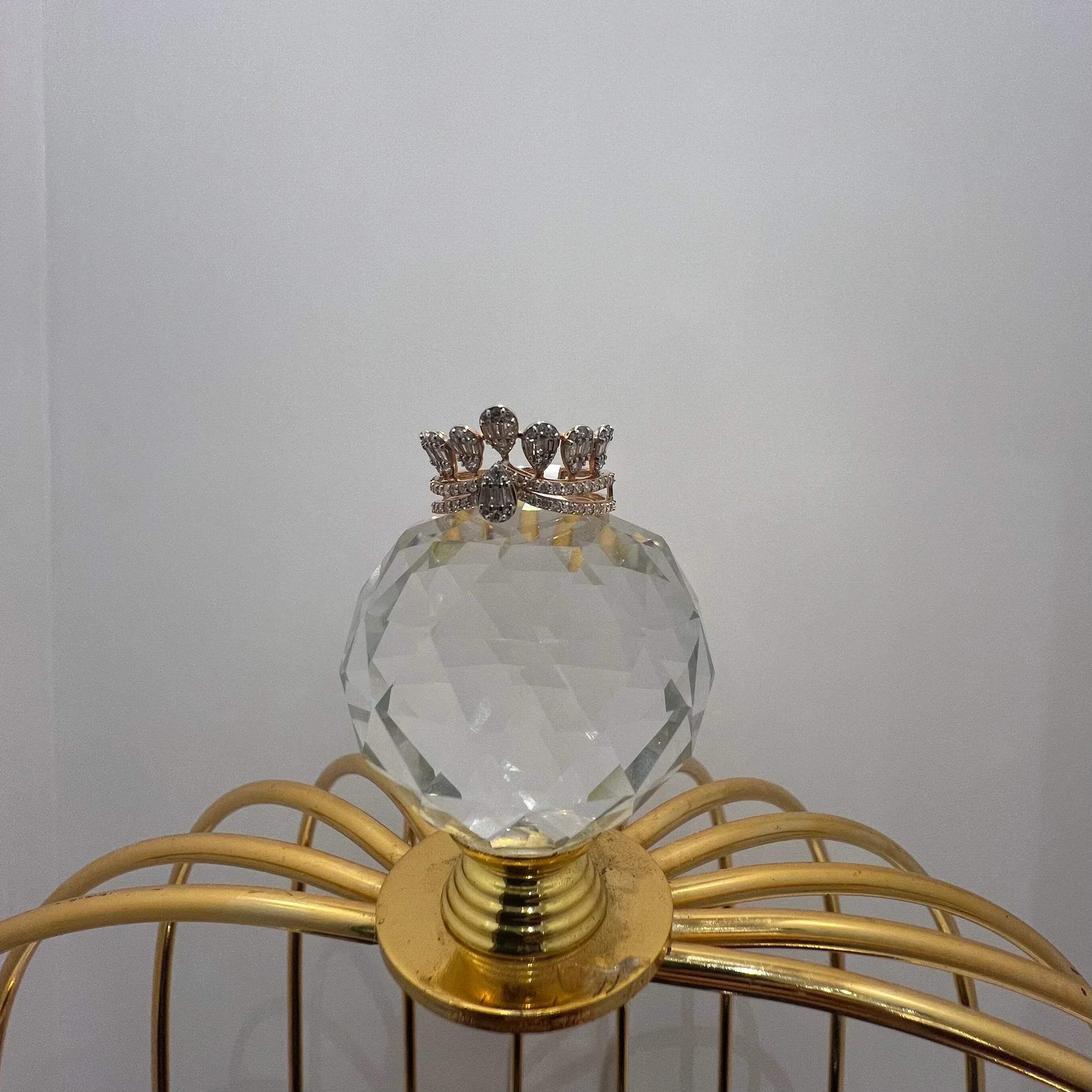 Close-up of a crown-inspired diamond ring featuring multiple baguette-shaped diamonds set upright along the top, paired with a pavé-set band in gold, displayed a glass ball that is on a gold color cage.