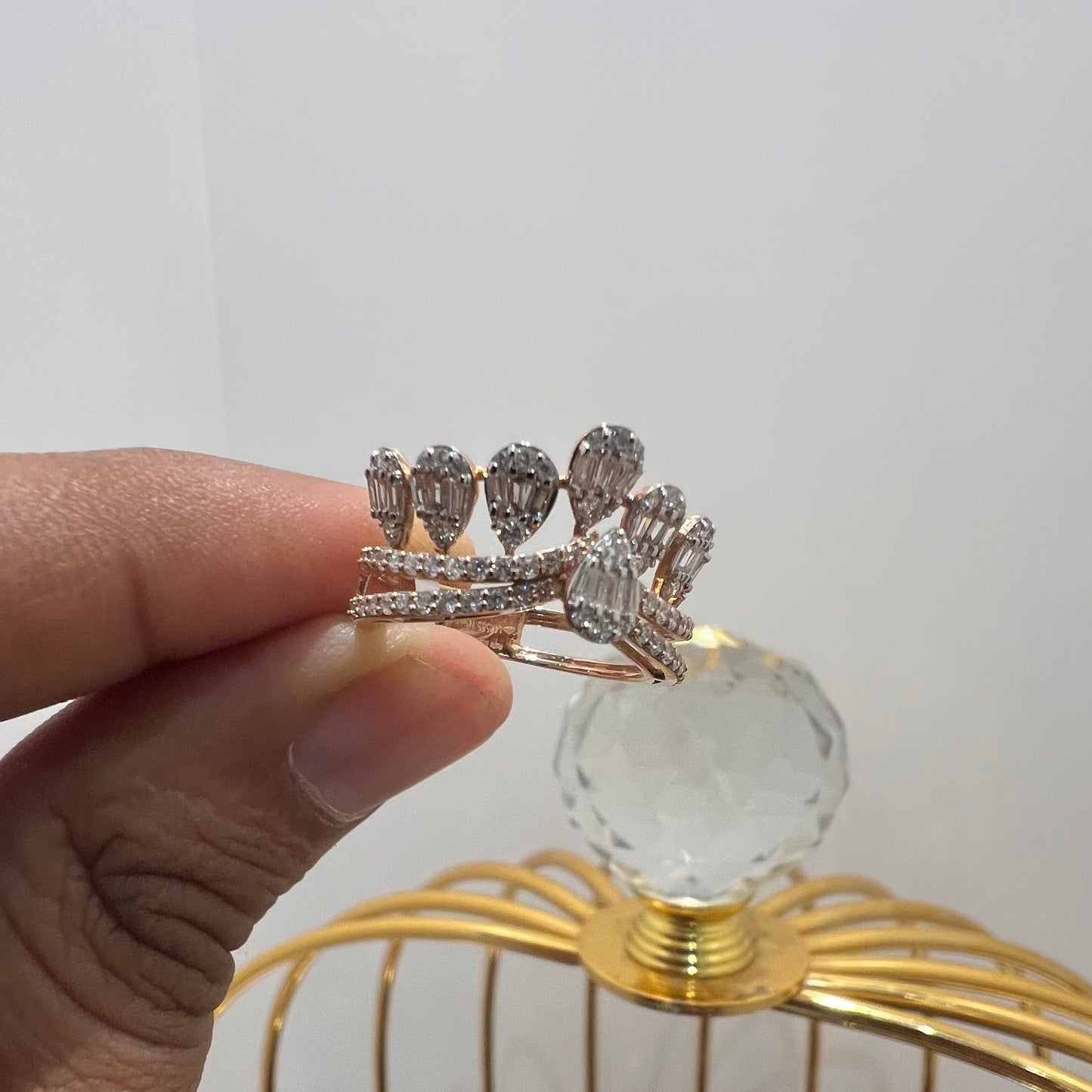 Close-up of a hand holding a crown-inspired diamond ring featuring multiple baguette-shaped diamonds set upright along the top, paired with a pavé-set band in gold, displayed against a gold color cage.