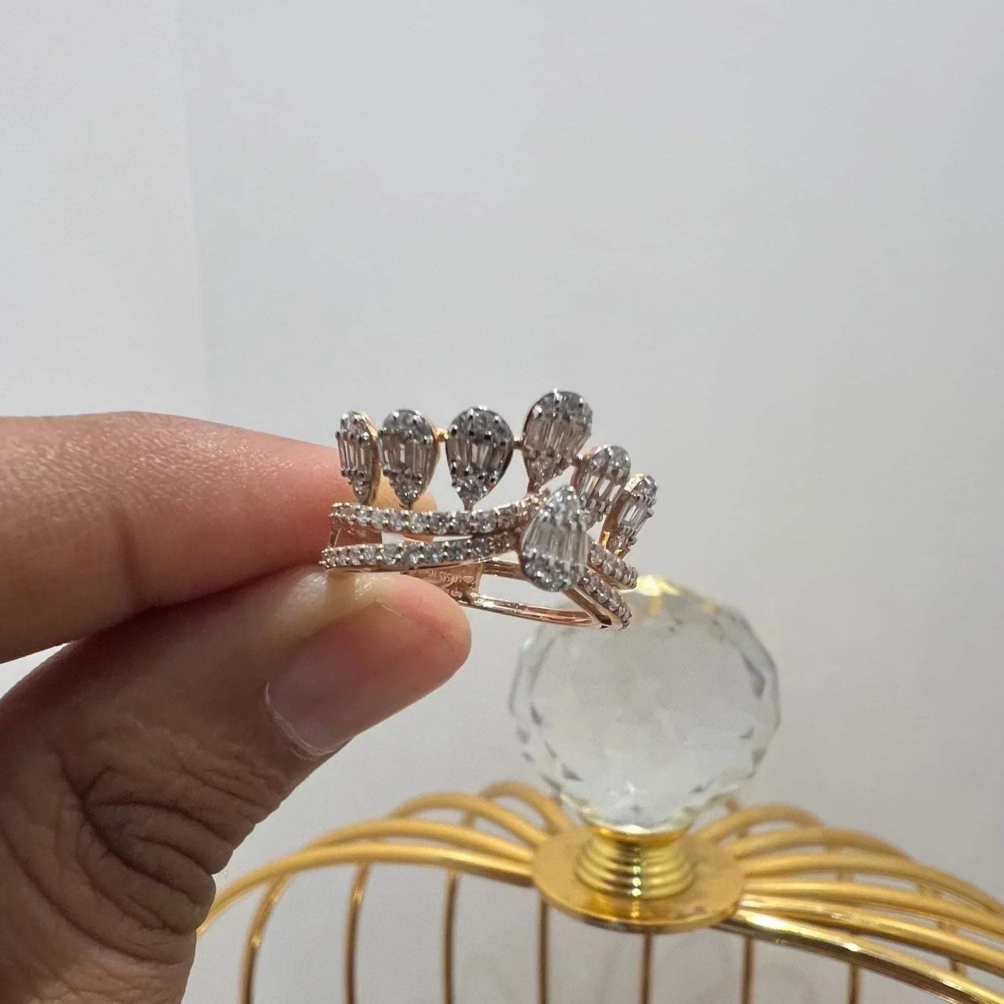 Close-up of a hand holding a crown-inspired diamond ring featuring multiple baguette-shaped diamonds set upright along the top, paired with a pavé-set band in gold, displayed against a gold color cage.