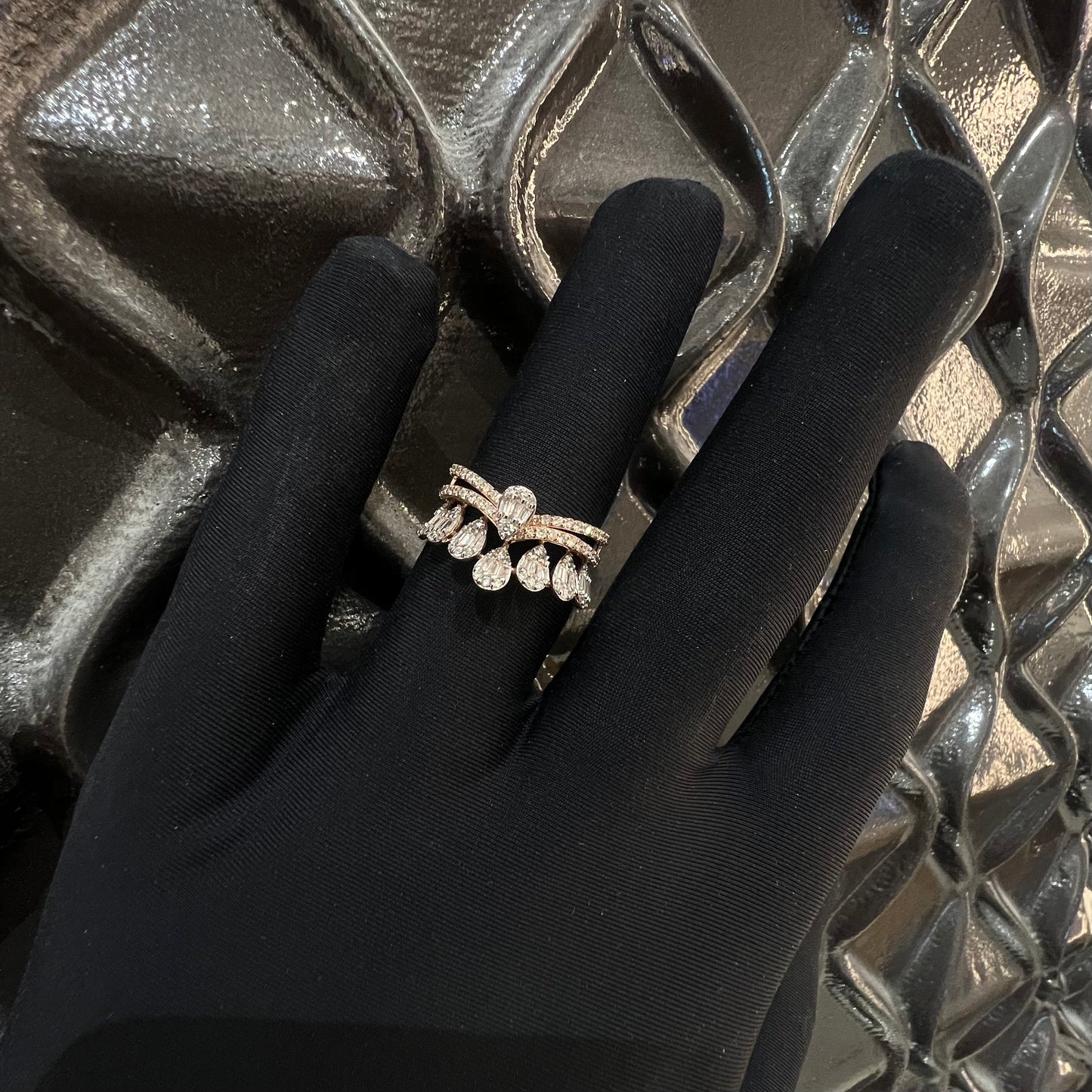 Close-up of a hand holding a crown-inspired diamond ring featuring multiple baguette-shaped diamonds set upright along the top, paired with a pavé-set band in gold, displayed against a light background with a decorative element.
