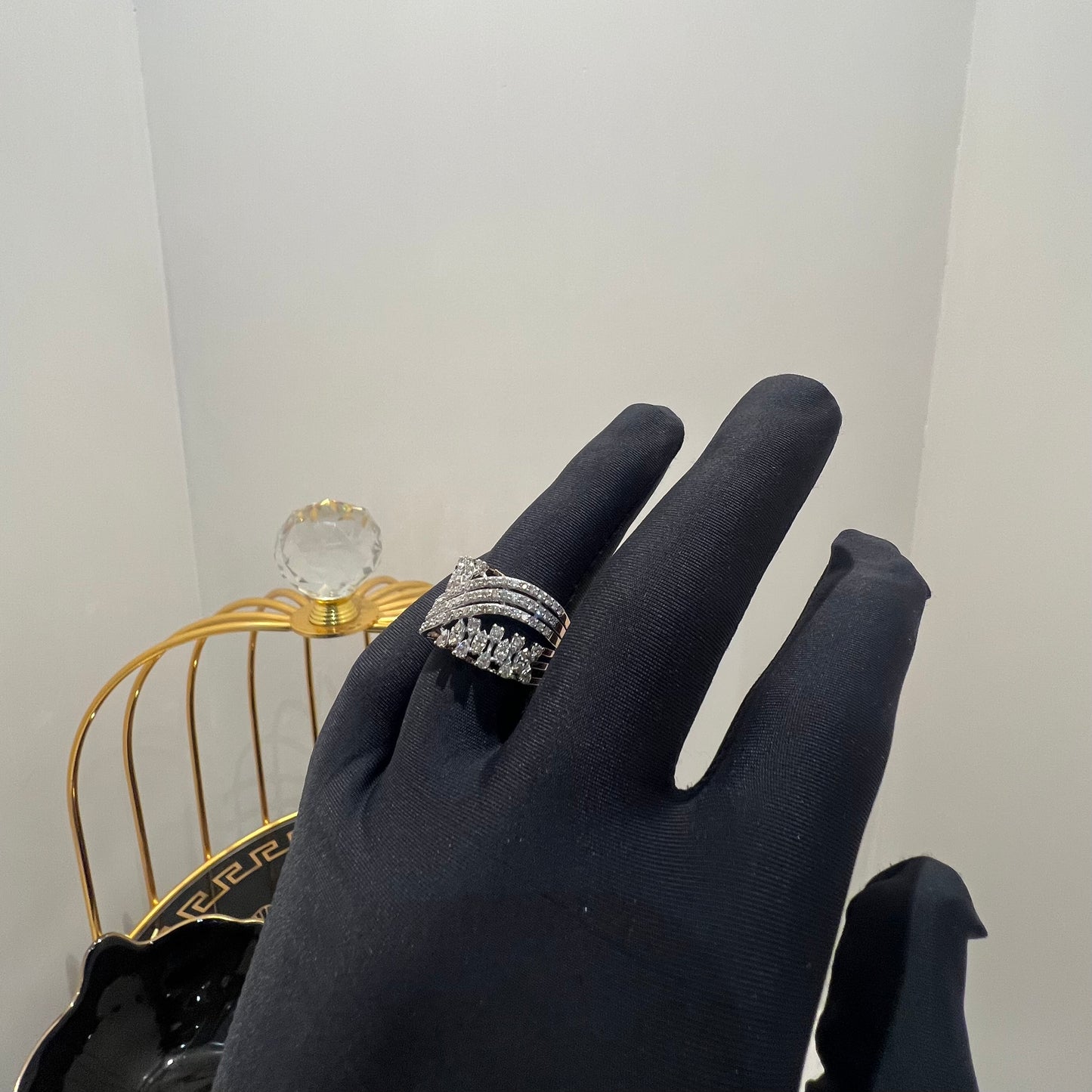 Close-up of an elegant wide diamond ring held by a gloved hand, featuring multiple crisscrossing bands adorned with round-cut diamonds set in gold, displayed against a gold decorative cage background.