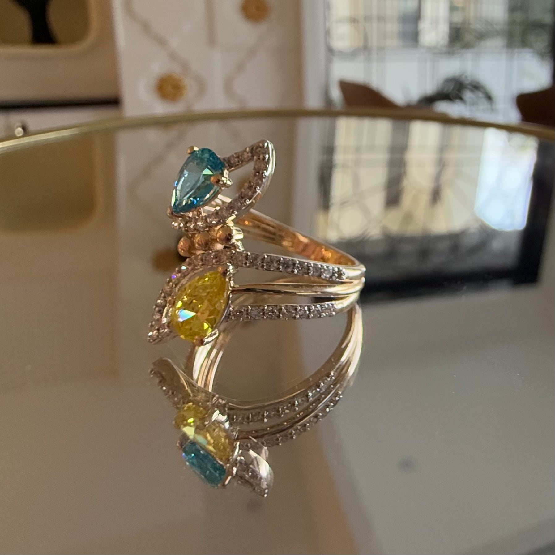 A reflective table displaying a two-stone diamond ring with yellow and blue colors, made of 14kt gold.